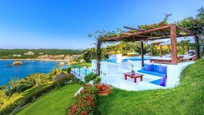 A luxurious outdoor pool area with a pergola, seating, and a view of the sea and buildings in the background. - Villa Roxana (Santa María Huatulco)