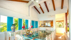 A dining area with a glass table, blue shutters, and a view of greenery outside. - Villa Roxana (Santa María Huatulco)