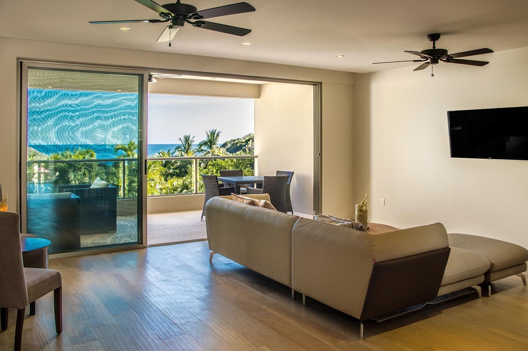 A modern living room with a sofa, a dining area with chairs, a TV mounted on the wall, and a balcony with a view of the ocean.