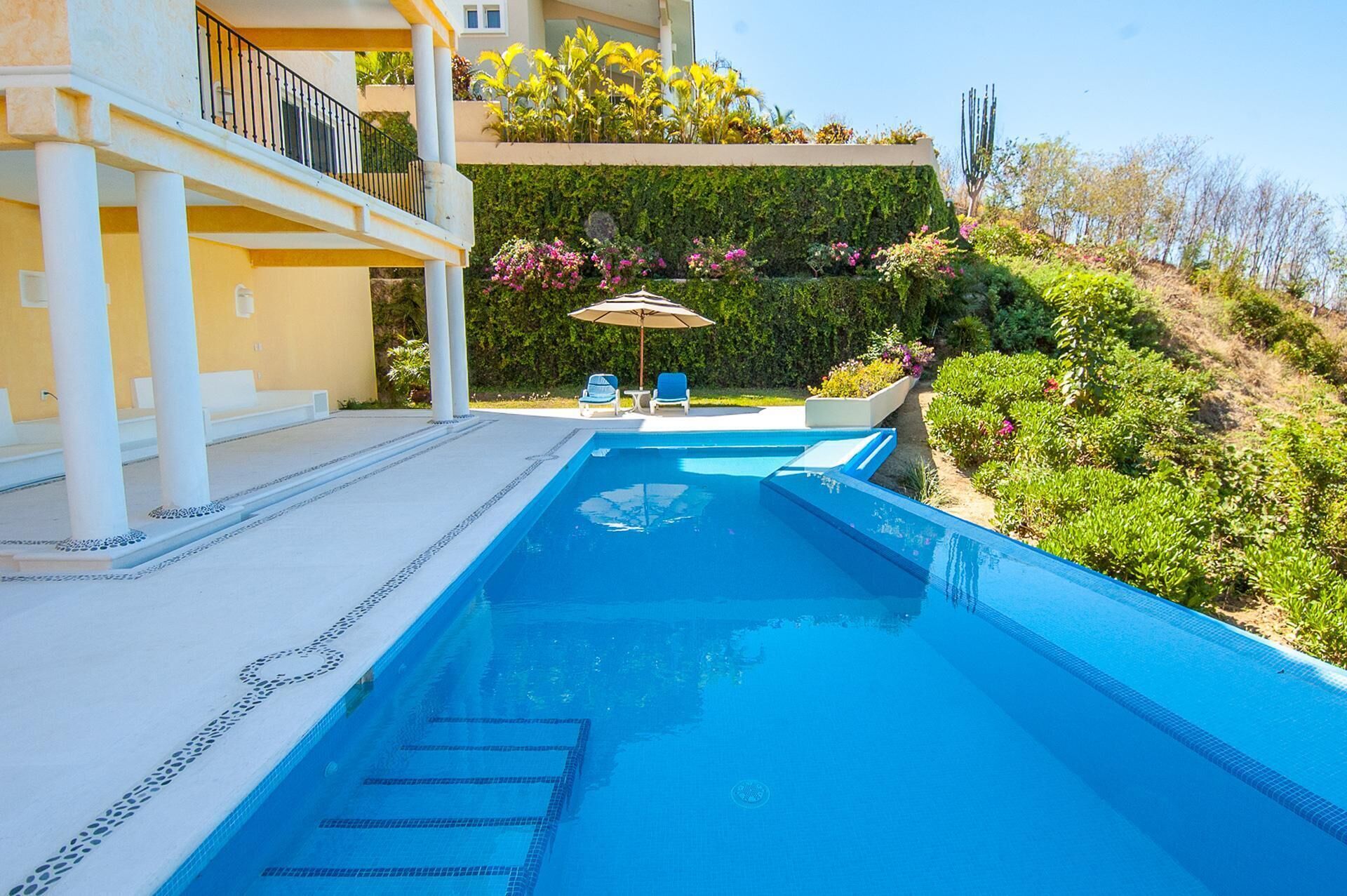 A swimming pool with a white tiled edge, a covered seating area with chairs and an umbrella, and a lush green garden with various plants and flowers.