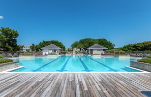 Une piscine rectangulaire à l’eau d’un bleu limpide, entourée d’une terrasse en bois. On y trouve des chaises longues et deux petits bâtiments dotés de toits inclinés.