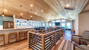 A spacious kitchen with wooden ceiling and bar stools, a dining area with a view, and a cozy seating area with patterned cushions. - Aftermath - Corolla, Ocean Sands, Oceanfront Luxury, Pool, Hot Tub, Elevator, Rec Room (Corolla)
