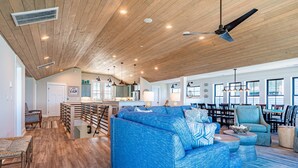 A spacious living area with a large blue sofa, wooden ceiling, and a dining area with chairs and tables. - Aftermath - Corolla, Ocean Sands, Oceanfront Luxury, Pool, Hot Tub, Elevator, Rec Room (Corolla)
