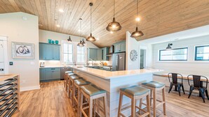 A modern kitchen with a central island, bar stools, and wooden flooring. - Aftermath - Corolla, Ocean Sands, Oceanfront Luxury, Pool, Hot Tub, Elevator, Rec Room (Corolla)