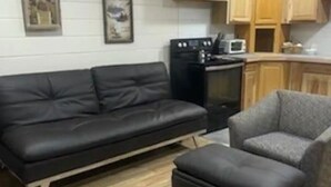 A living room with a black sofa, a grey armchair, and a small ottoman. There is a kitchen area with wooden cabinets and a microwave. - Captain's Quarters (Bayview)