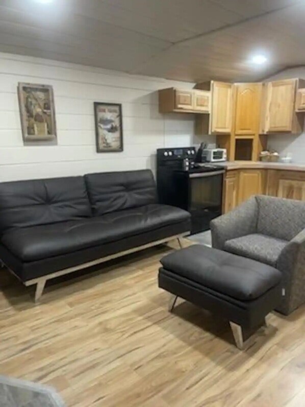 A living room with a black sofa, a grey armchair, and a small ottoman. There is a kitchen area with wooden cabinets and a microwave. - Captain's Quarters (Bayview)