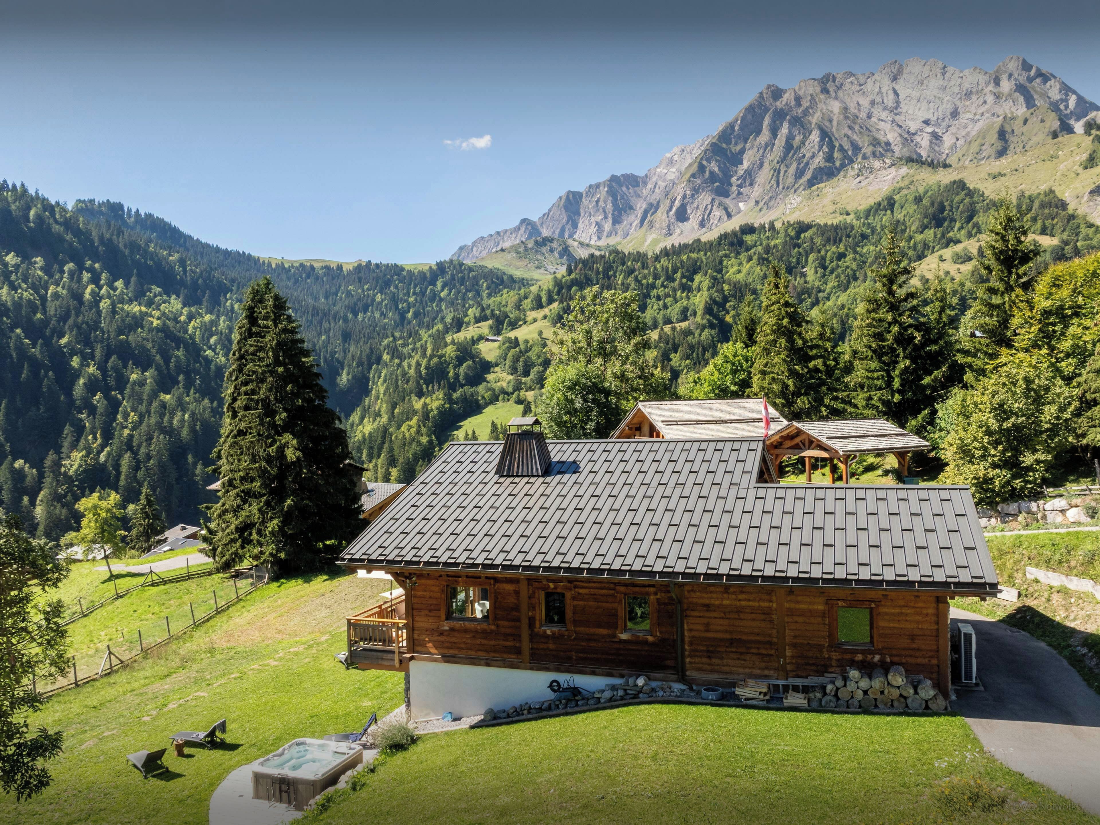 Une cabane en bois, dotée d’un toit en pente, entourée d’un paysage verdoyant et luxuriant, avec des montagnes en arrière-plan.