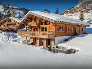 A wooden chalet with a balcony, stone foundation, and a hot tub in the snow. - Chalet Sabaroc - modern ski base for 10, hot tub & views - OVO Network (La Giettaz)