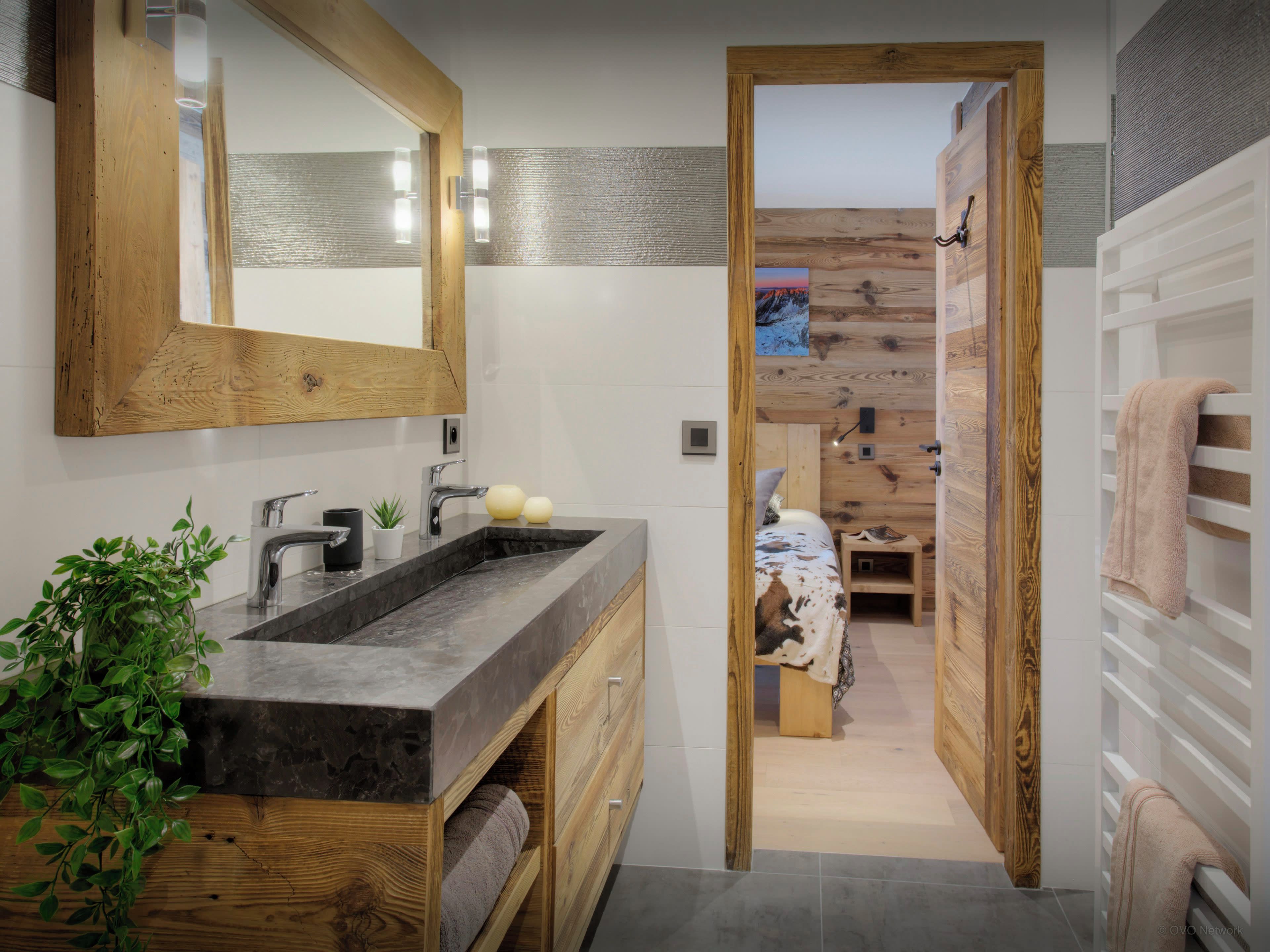 Un baño moderno con tocador de madera, un espejo grande y vistas a un dormitorio con pared de madera y una cama.