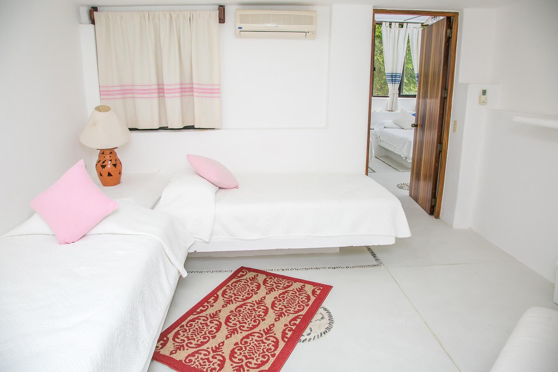 A minimalist bedroom with two beds, a red patterned rug, a lamp, and a window with curtains.