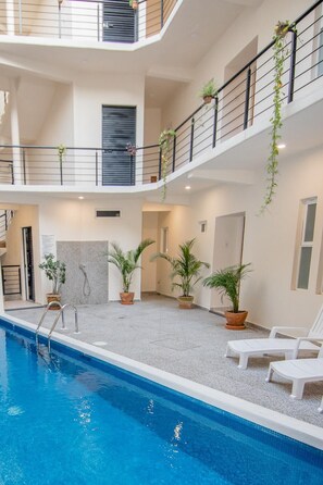 Un área de piscina interior con varios niveles, piscina cristalina, plantas en macetas y sillones blancos para relajarse.