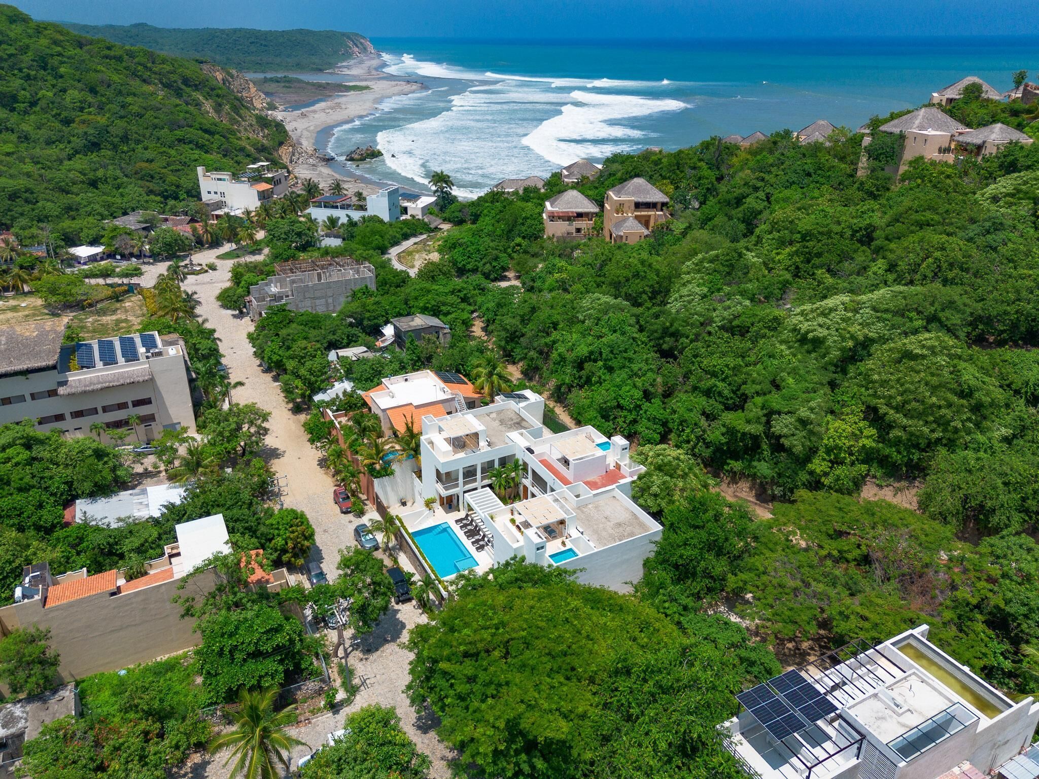 An aerial view of a coastal resort with multiple buildings, a swimming pool, and lush greenery.