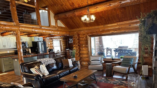 Interno di una baita di legno con divano in pelle, zona pranzo, cucina e ampie finestre.