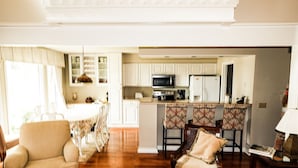 A spacious living room with a high ceiling, a dining area with patterned chairs, and a kitchen with white cabinets and modern appliances.