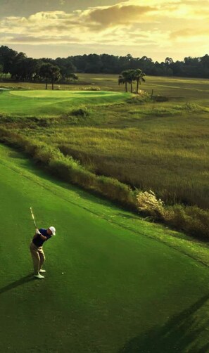 Un campo de golf con un jugador de camisa azul y gorra blanca preparándose para golpear la pelota.