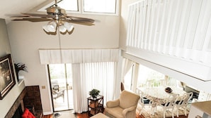 A spacious living room with a high ceiling fan, a fireplace, a dining area with a table and chairs, and a large window allowing natural light.