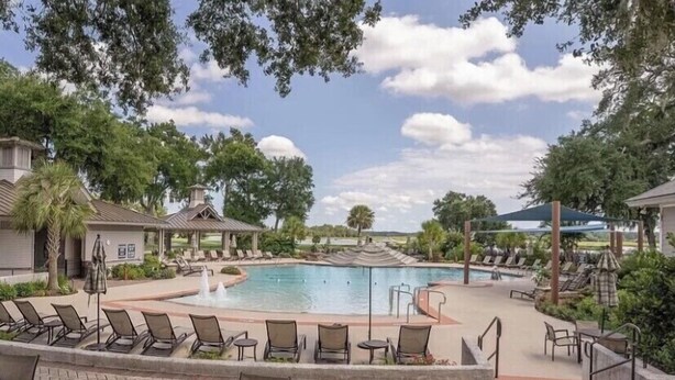 A pool area with lounge chairs and tables, surrounded by trees and a pavilion.