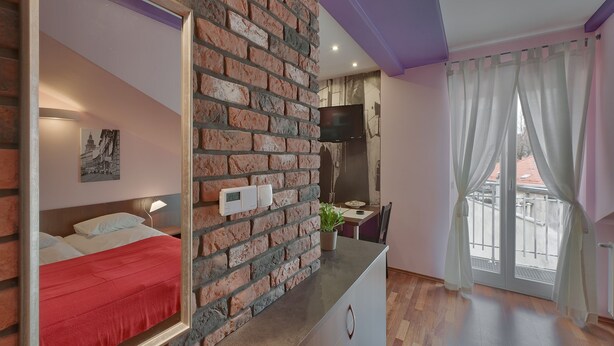 Ein Hotelzimmer mit Ziegelwand, einem Bett mit roter Decke, einem an der Wand befestigten Fernseher, einem kleinen Tisch mit Pflanze und einem Balkon mit Blick.