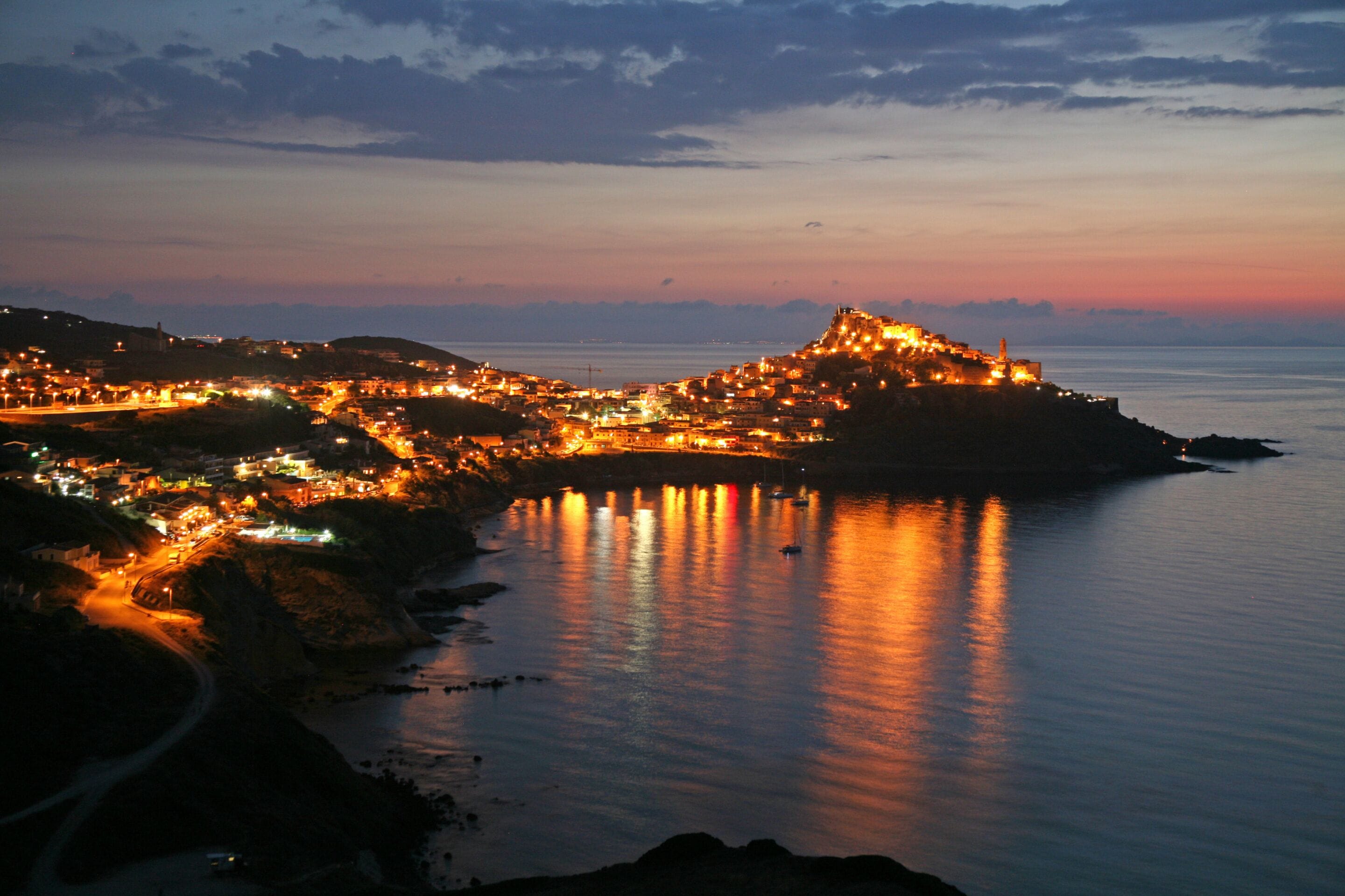 Une ville côtière au crépuscule, avec des bâtiments illuminés et une structure imposante qui se dresse sur une colline.