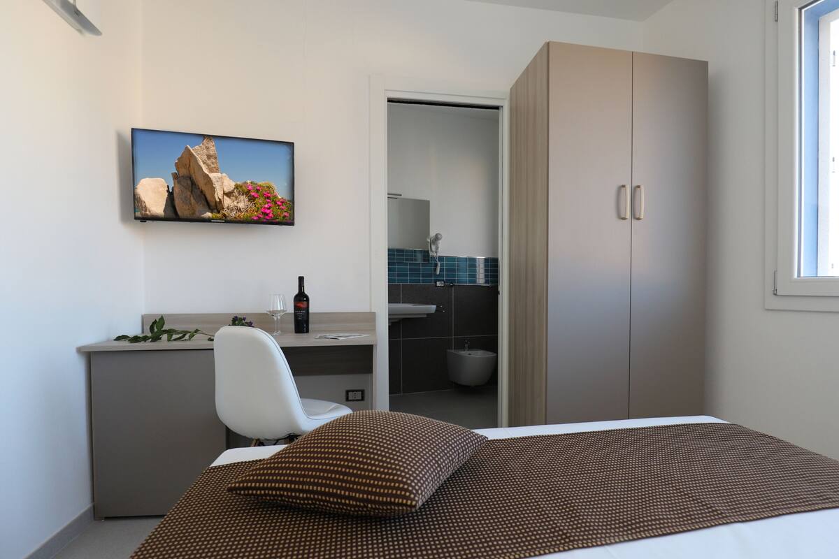 Un dormitorio moderno con una cama, un escritorio, una silla, un armario y un baño visible a través de una puerta abierta.