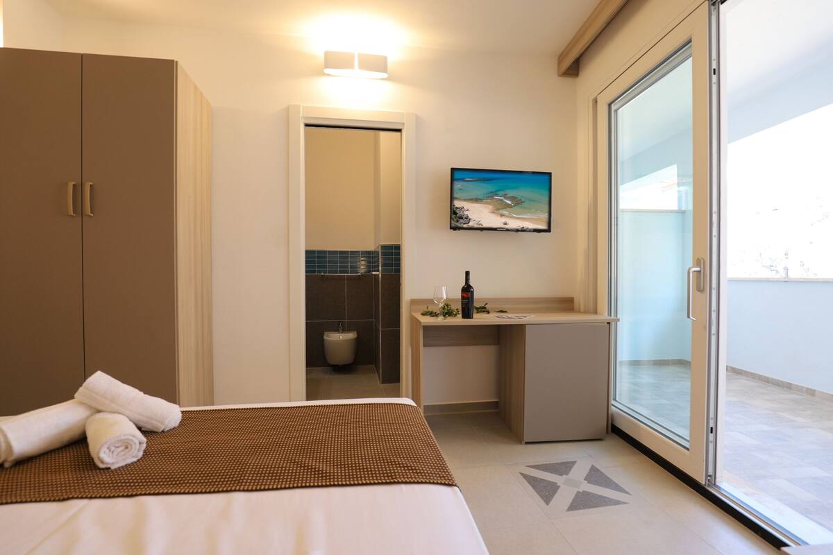 Una habitación de hotel con cama, escritorio, baño y puerta corrediza de vidrio al exterior.