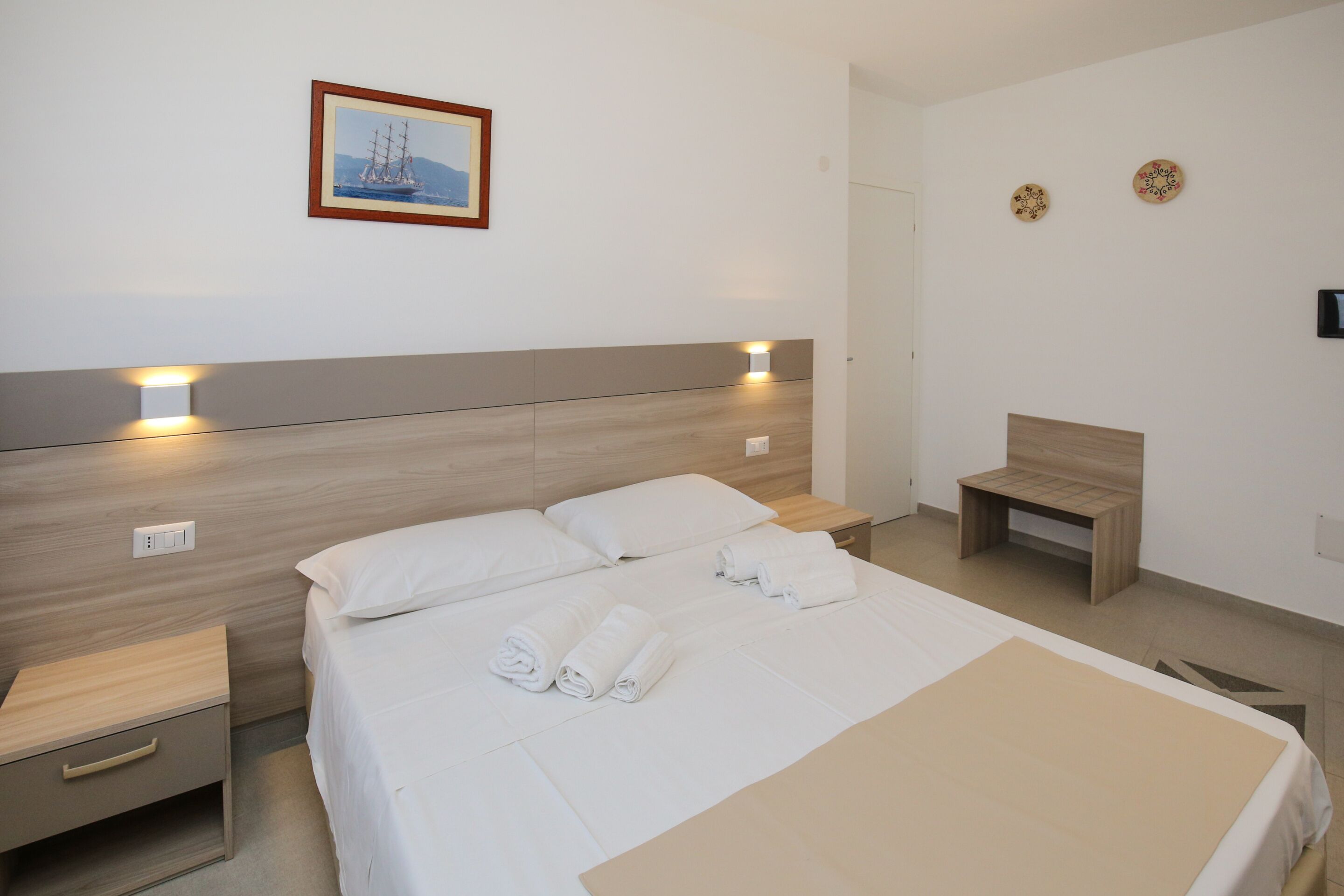 Una habitación de hotel moderna con cabecera de madera, cama con ropa de cama blanca, mesita de noche con lámpara y un pequeño banco de madera.