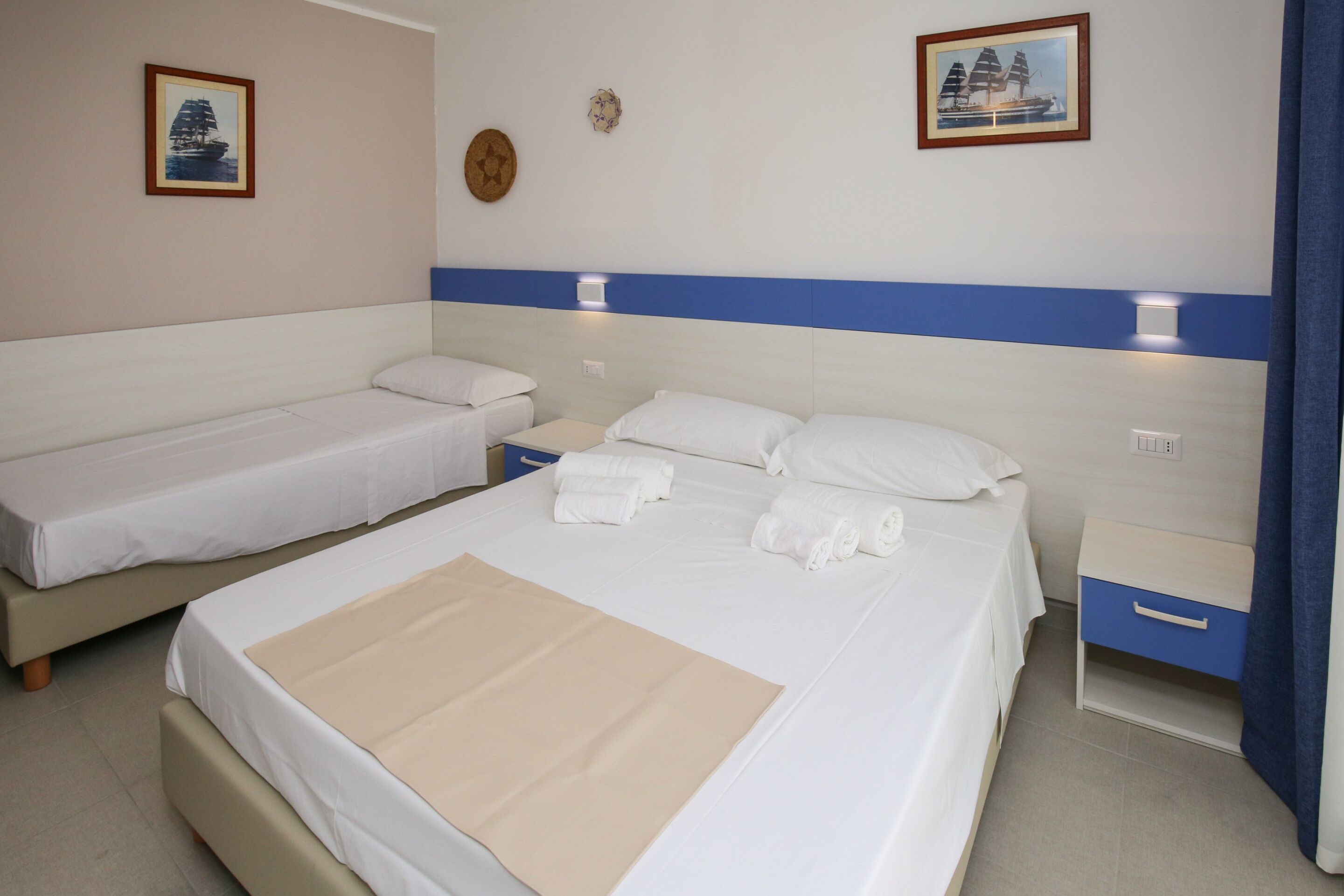 Habitación de hotel con dos camas individuales, una cama más grande con una colcha color beige, una mesita de noche con una lámpara y un pequeño mueble con cajones.