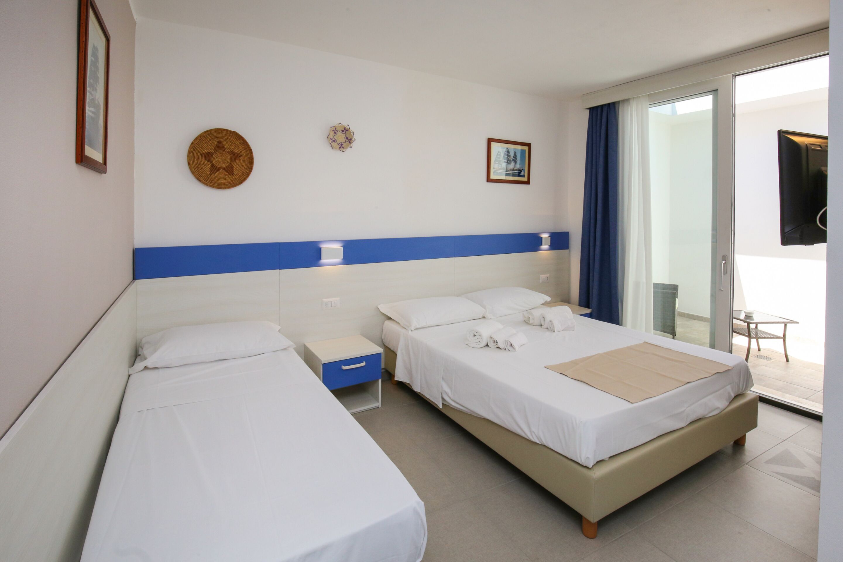 A hotel room with two single beds, a small table, a chair, and a large sliding glass door leading to an outdoor area.