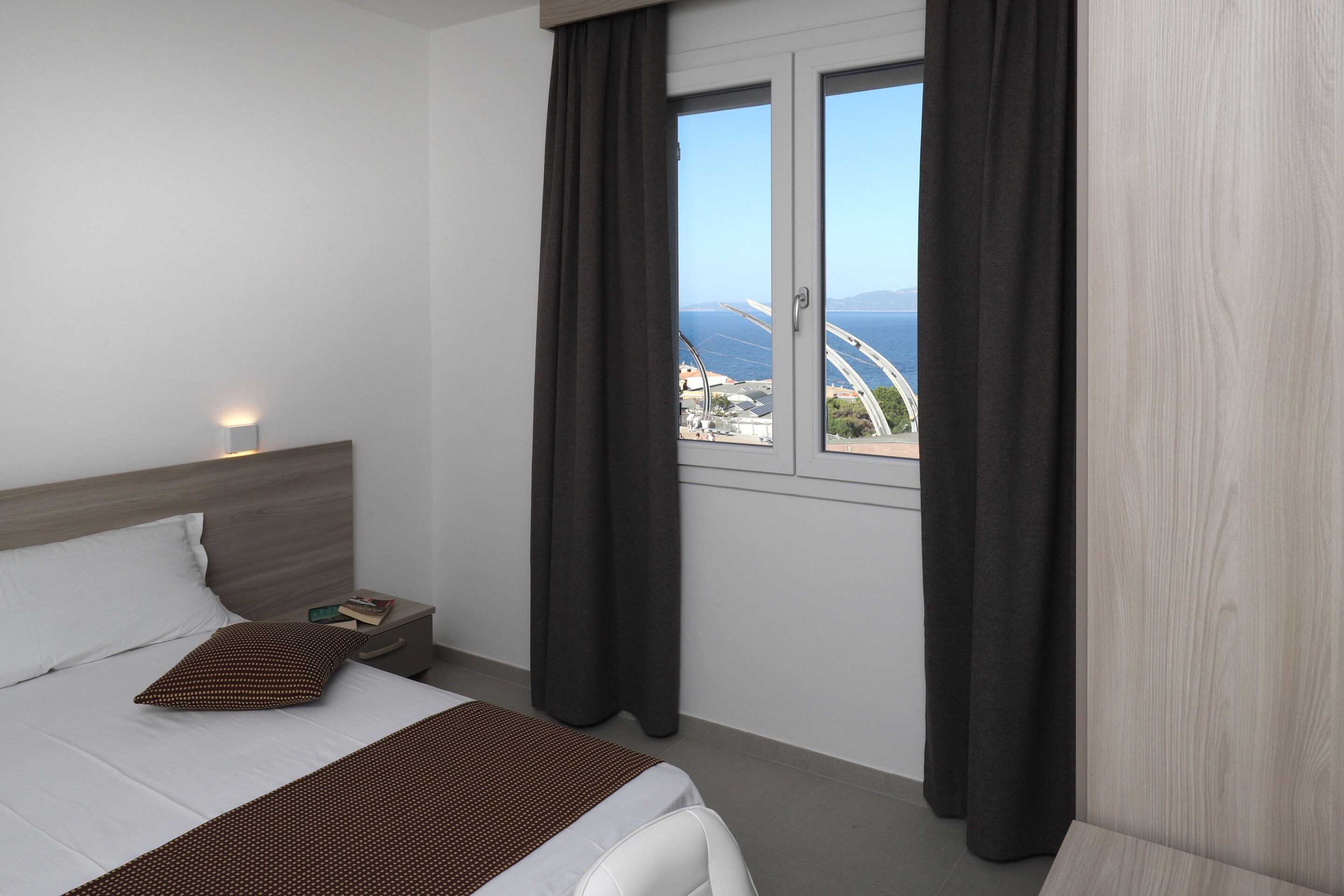 Habitación de hotel con cama, mesita de noche, ventana con cortinas y vista a un puente y paisaje urbano.