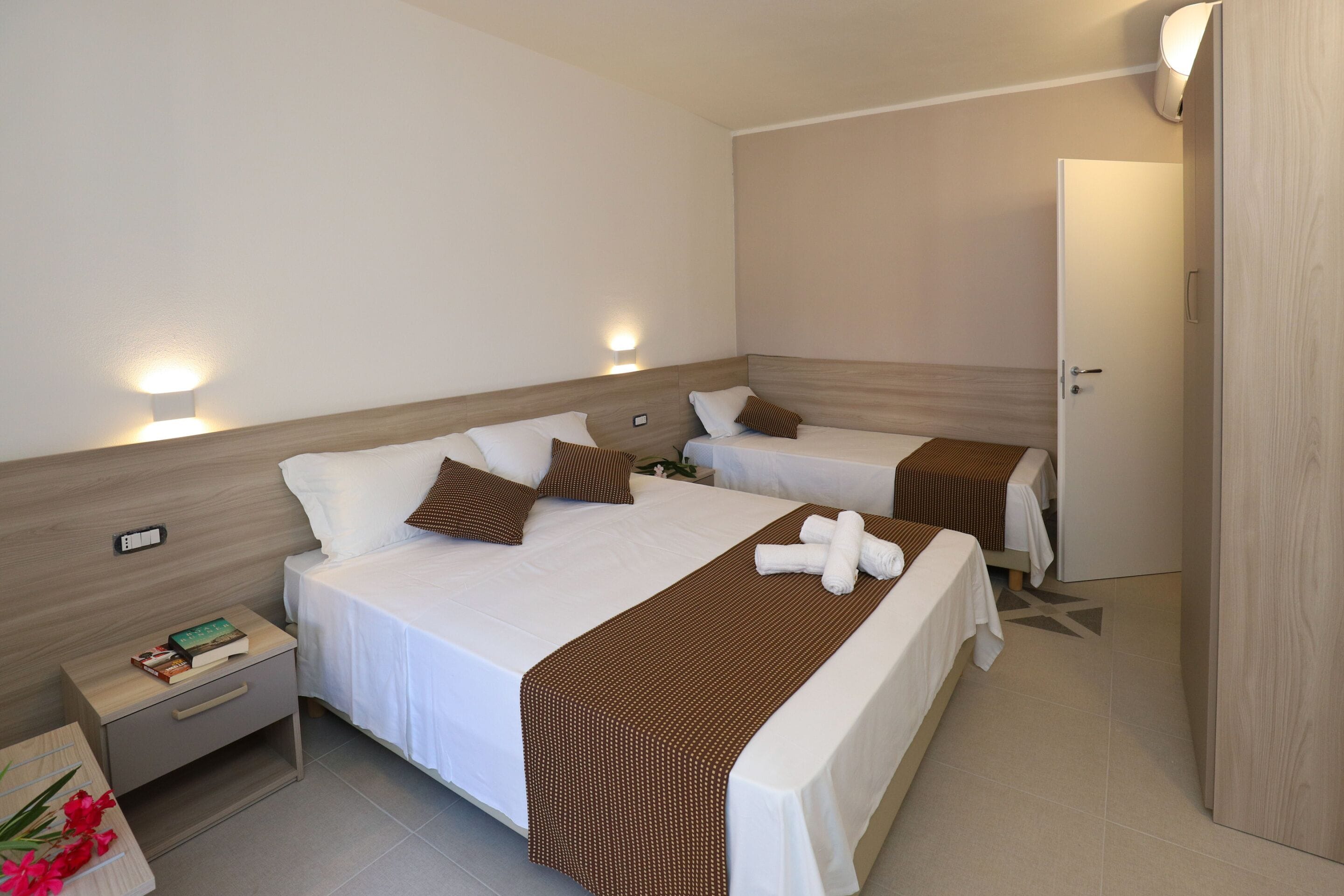 A hotel room with two beds, a nightstand with a book, a wall-mounted light, and a door.