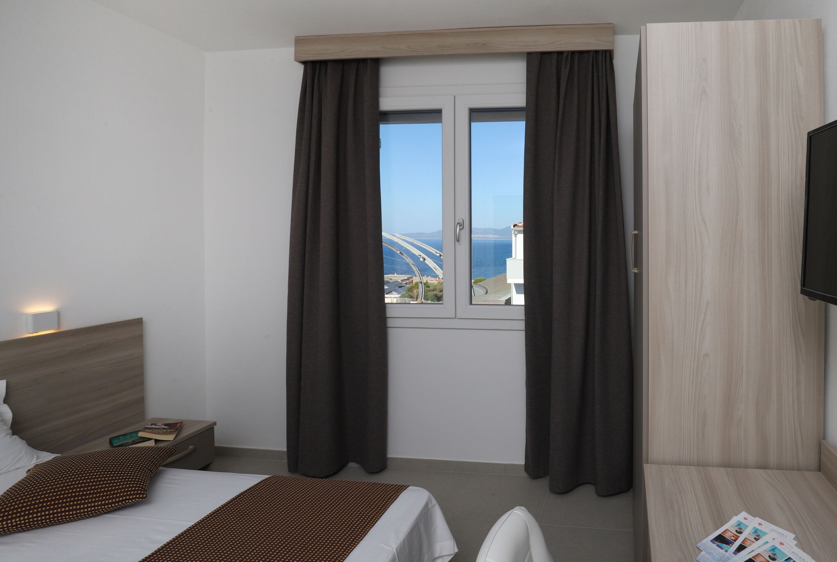 A hotel room with a bed, a nightstand, a TV, a window with curtains, and a view of a bridge and sea.