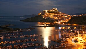 Un poble costaner de nit amb un castell imponent sobre una collada, un port esportiu amb vaixells i un pont que uneix el poble al continent.