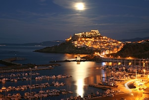 A coastal town at night with a prominent castle on a hill, a marina with boats, and a bridge connecting the town to the mainland.
