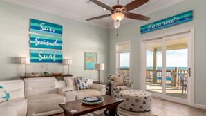 A living room with a ceiling fan, a sign reading "Encantado Beach House," and a view of the ocean through a sliding glass door. - Encantado | Cancellation 9/26 week. Pool! (Dauphin Island)