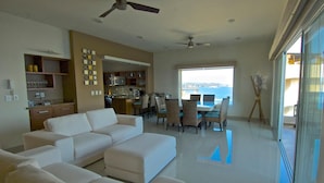 A spacious living room with a large white sofa, a dining area with wicker chairs, a kitchen with modern appliances, and a view of the sea through large windows. - Punta Arrocito PH (Santa María Huatulco)