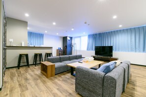 Una sala moderna con un sofá, una mesa de centro y un televisor. Hay dos taburetes y una pequeña área de cocina con mostrador y pileta.