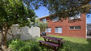 A backyard with a wooden picnic table, a brick building, and a white fence. - Scenic Beach Apartment (Apollo Bay)
