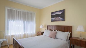 A bedroom with a bed, two bedside tables, a chair, a window with sheer curtains, and a painting on the wall. - Scenic Beach Apartment (Apollo Bay)