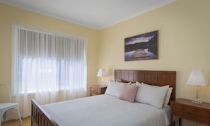 Ein Schlafzimmer mit einem Bett, zwei Nachttischen, einem Sessel, einem Fenster mit durchsichtigen Vorhängen und einem Bild an der Wand.