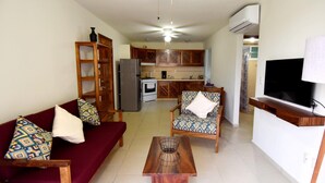 A living room with a sofa, armchair, and coffee table. There is a kitchen in the background with a refrigerator and stove. - Terrazas C7 (Santa María Huatulco)