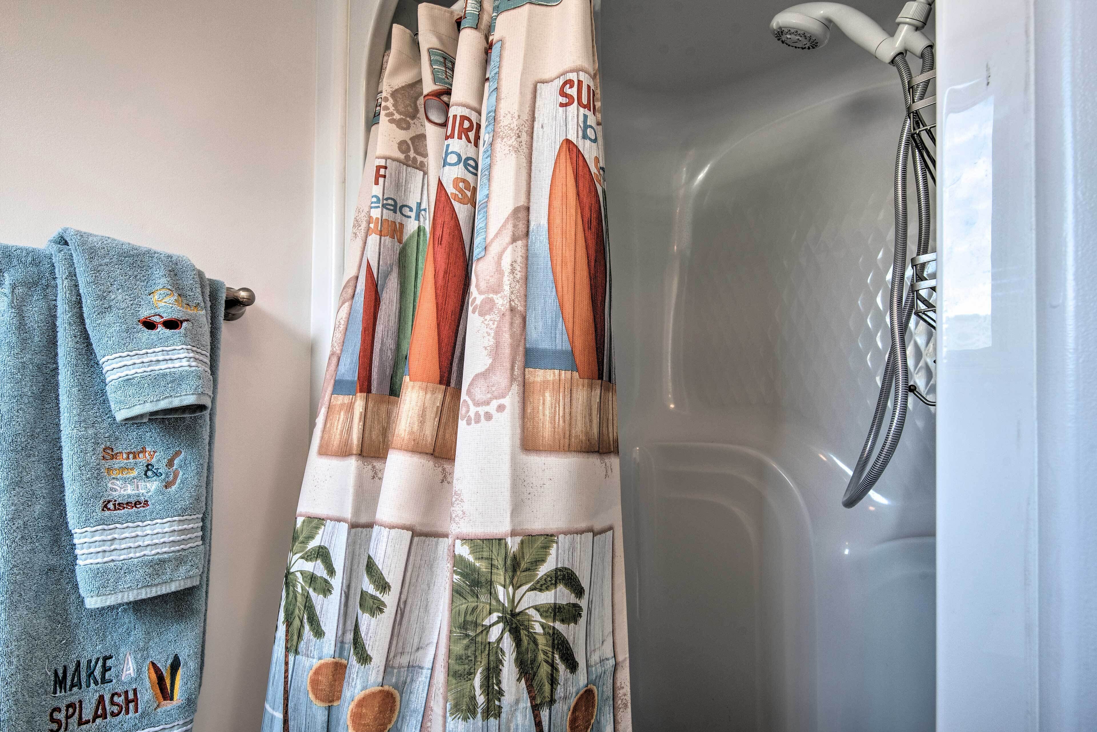 A bathroom with a shower curtain featuring palm trees and the text "Surf's Up." A towel with the text "Make a Splash" is hanging on the wall.