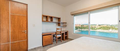 A bedroom with a large bed, a wooden desk with a chair, a wardrobe, and a large window offering a view of the sea and landscape.