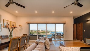 A spacious living room with a large window offering a view of the sea, a dining area with a wooden table and chairs, and a modern ceiling fan. - Punta Arrocito B202 (Santa María Huatulco)