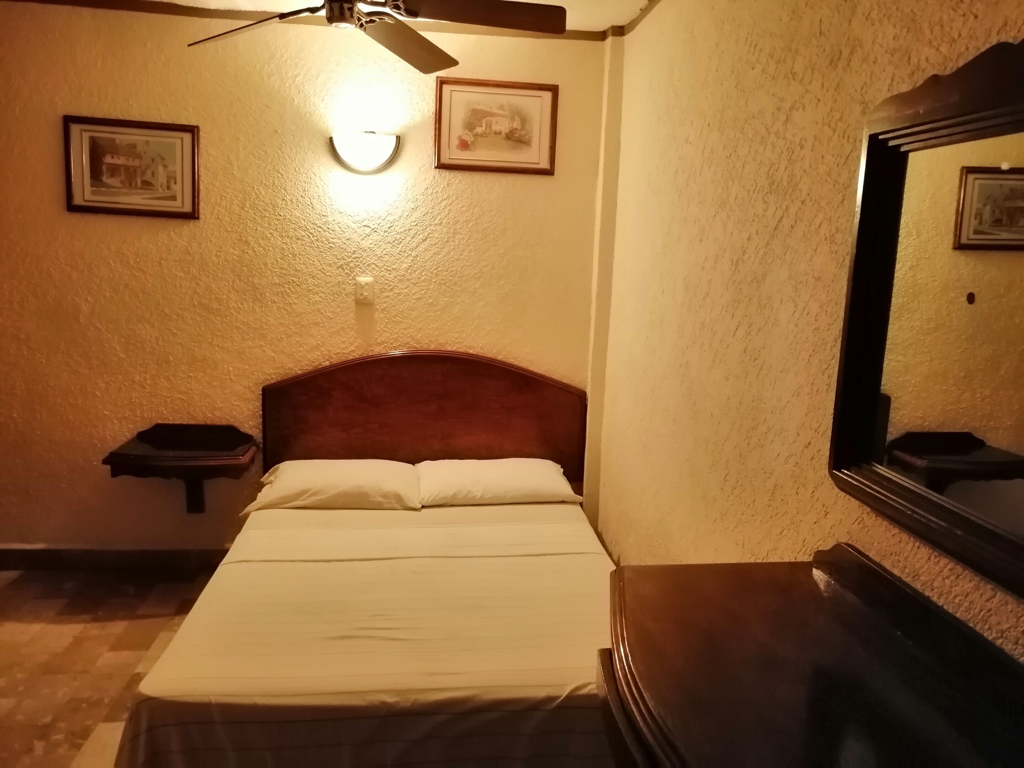 Une chambre d’hôtel équipée d’un lit, d’une table de chevet, d’un ventilateur de plafond et d’une lampe fixée au mur.