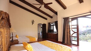 A bedroom with two beds, wooden headboard, a ceiling fan, a large window with curtains, and a view of the outside. - Room "El Guayabo" Caroline Bella  (Ajijic)