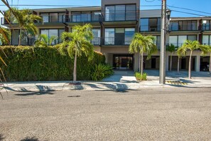 A modern multi-story building with balconies, surrounded by palm trees and greenery.