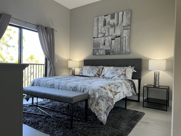 A modern bedroom with a large bed, two bedside lamps, a bench, a rug, and a view of greenery outside.