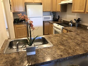 A kitchen with granite countertops, wooden cabinets, a stainless steel sink, a white refrigerator, a microwave, a stove, and a potted orchid.