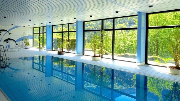 An indoor swimming pool with clear glass walls, potted plants, and a tiled floor. - Apartment 424 with indoor pool (Bad Reichenhall)