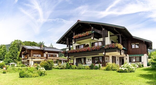 A large, two-story wooden building with balconies adorned with flowers, surrounded by lush greenery. - 2-room apartment, eat-in kitchen, sep. Living room and bedroom, large bathroom and infrared cabin (Reit im Winkl)